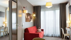 Habitación de hotel con una mesa de comedor preparada para dos personas, un sofá rojo, un ventanal con cortinas y un cuadro en la pared.