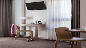 Habitación de hotel con televisión de pantalla plana montada en la pared, un escritorio con lámpara, una silla, una mesa con vaso y una ventana con cortinas.