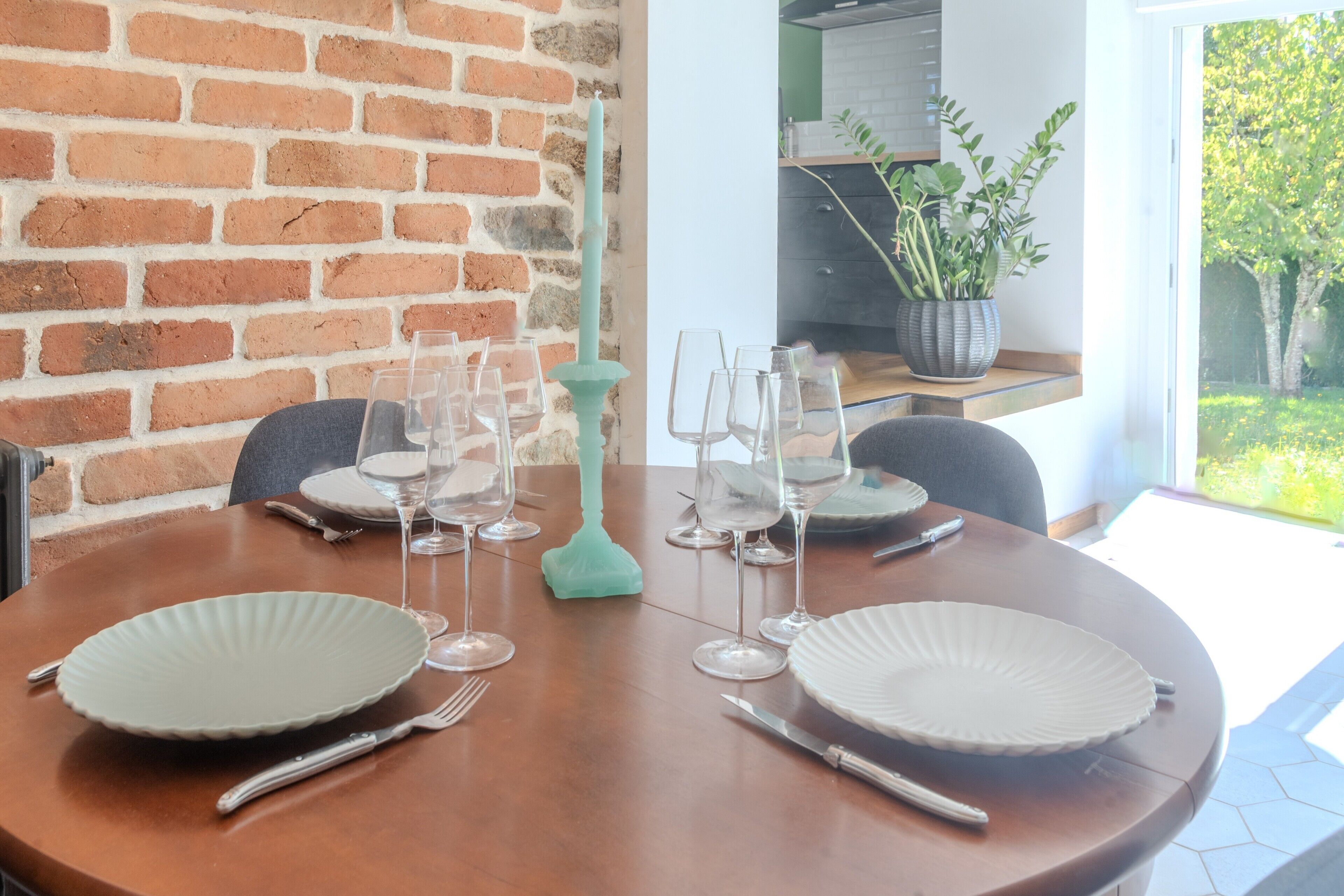 Une table à manger dressée avec des assiettes, des couverts et des verres à vin, un centre de table composé d’une bougie turquoise et un mur en briques.