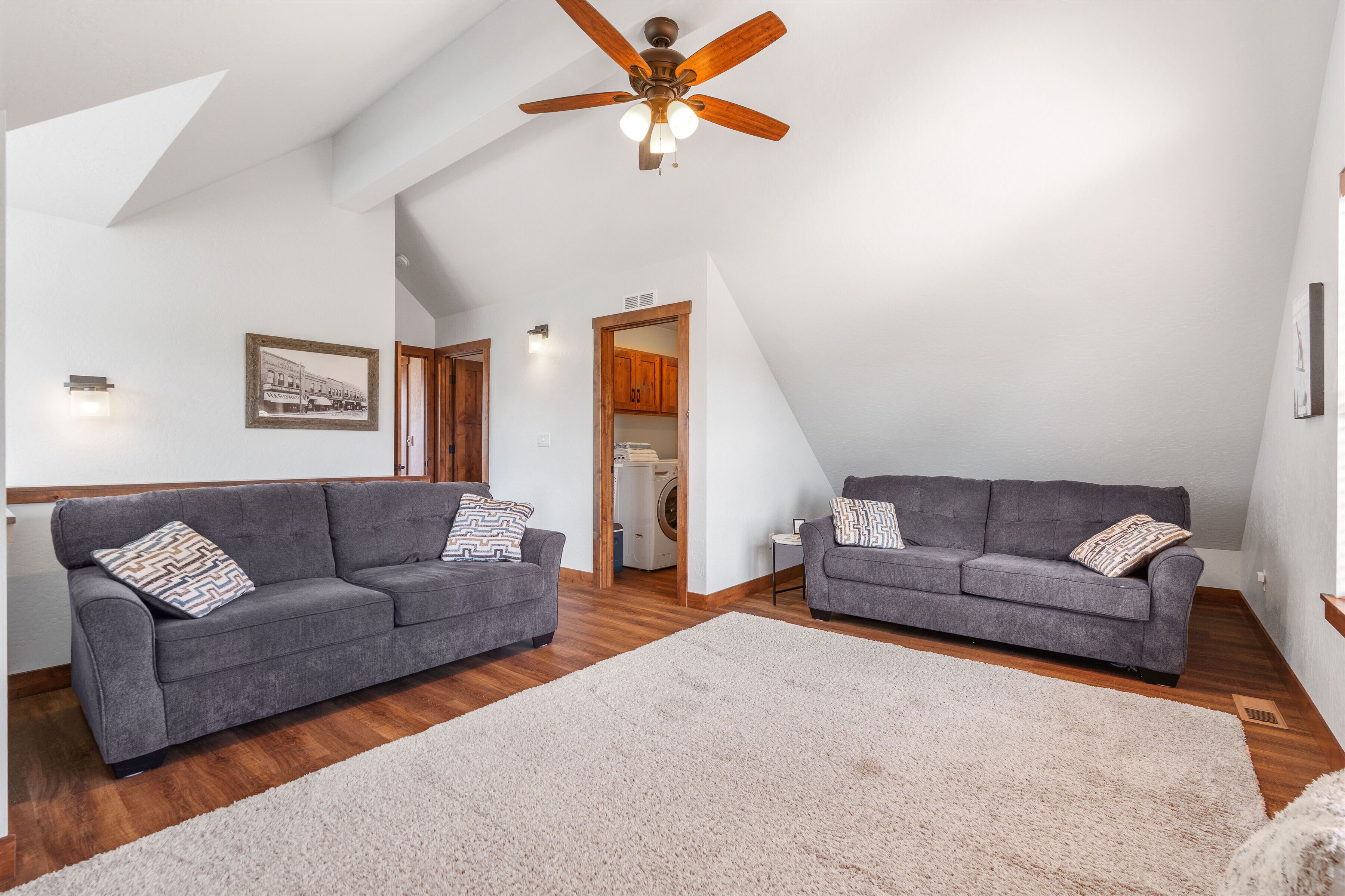 A spacious living room with two grey sofas, a large white rug, wooden flooring, a ceiling fan, and a framed picture on the wall.