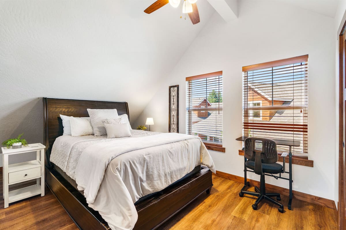 A bedroom with a large bed, a nightstand, a desk with a chair, and a window with blinds.
