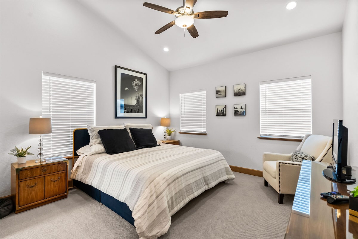 A bedroom with a bed, nightstands, a desk, a chair, a ceiling fan, and wall-mounted pictures.