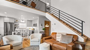Ein modernes Wohnzimmer mit einem braunen Ledersofa, zwei weißen Sesseln und einem Couchtisch. Eine Treppe mit schwarzer Geländer führt auf eine obere Ebene.