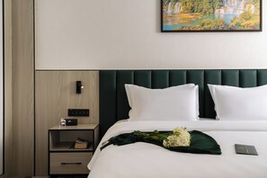 Ein Hotelzimmer mit einem ordentlich bezogenen Bett, einem Nachttisch mit Lampe, einem Gemälde an der Wand und einem weiteren Nachttisch mit Buch und Flasche.