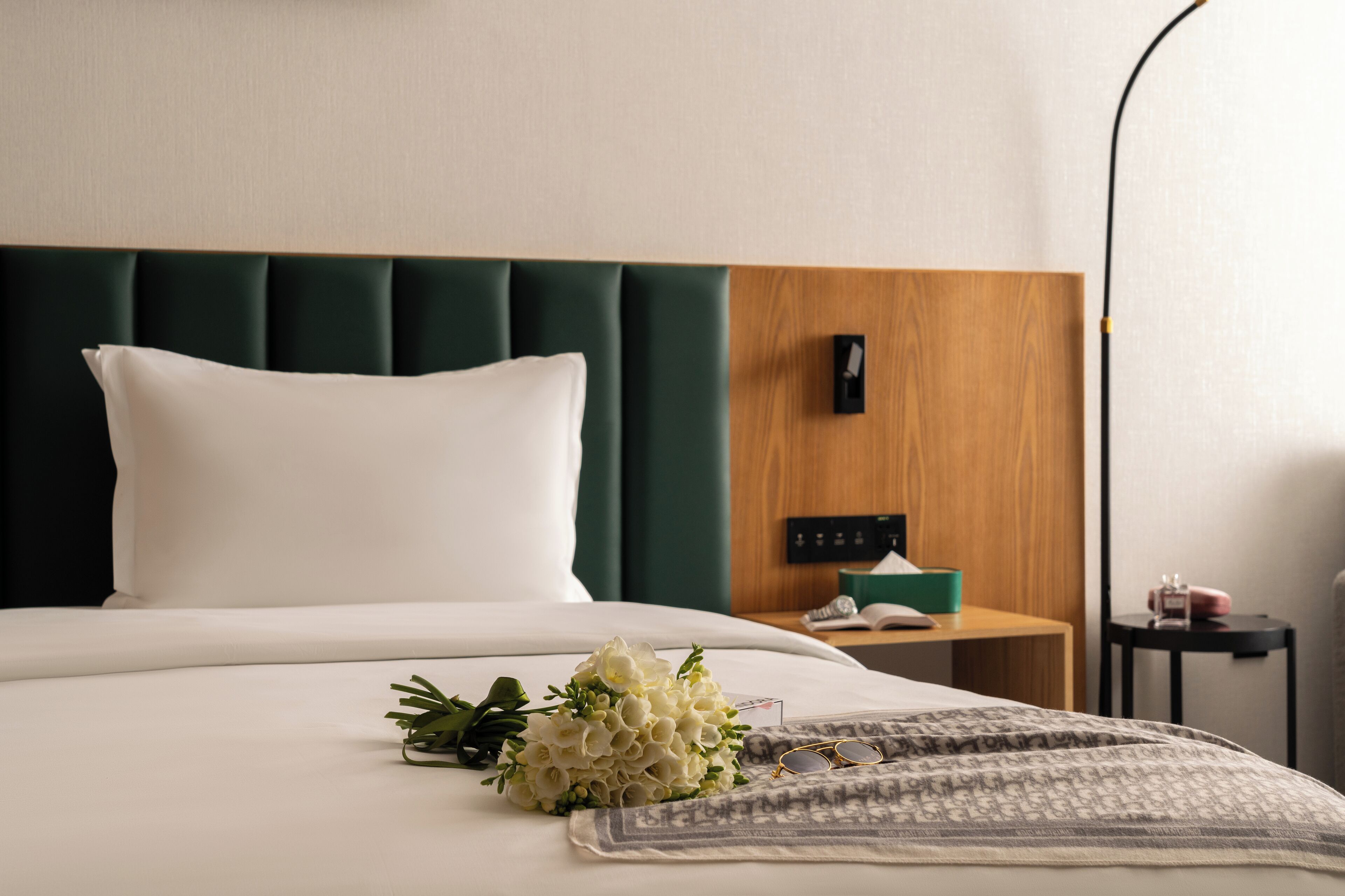 A neatly made bed with a green upholstered headboard, a bedside table with a lamp, and a small nightstand with a clock.