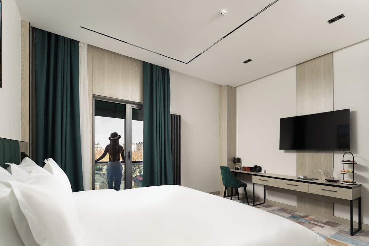 A modern hotel room with a large bed, a desk with a flat-screen TV, and a view of a cityscape through a large window.