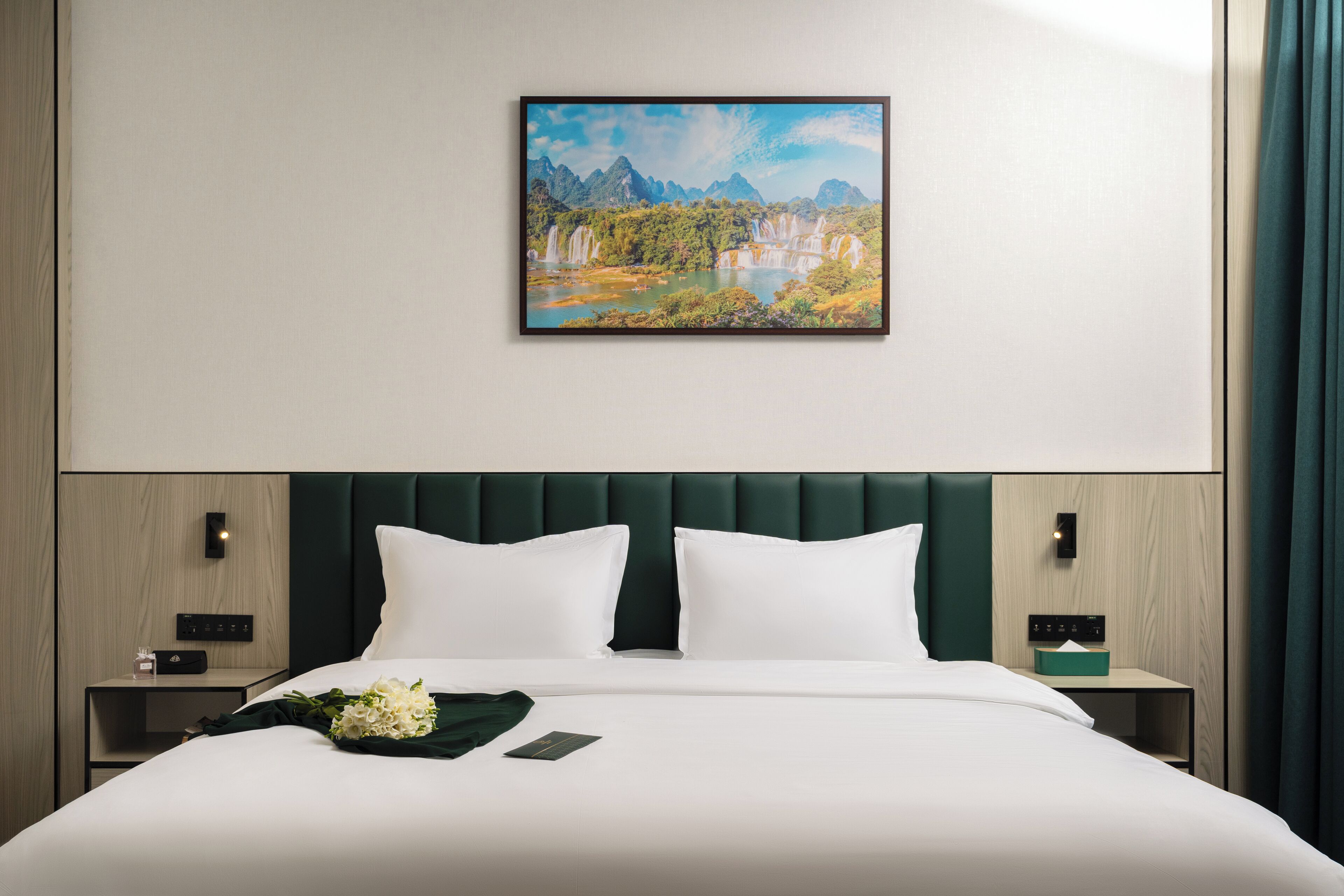 Una habitación de hotel con una cama grande, cabecera, mesita de noche, lámpara de pared y un cuadro enmarcado con un paisaje.