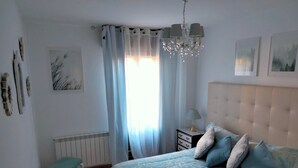 Une chambre à coucher avec un lit, une table de chevet, une chaise, un radiateur et une fenêtre avec des rideaux.