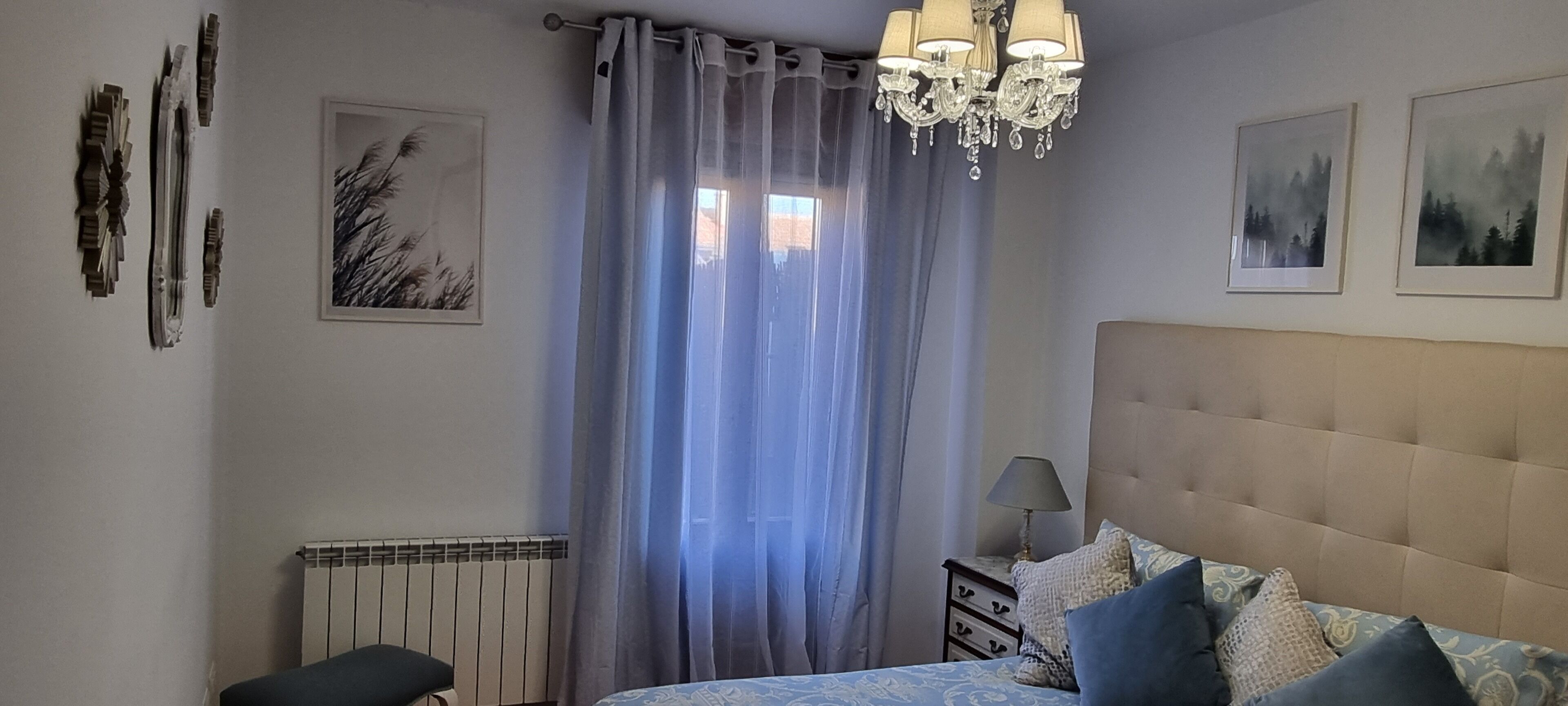 Un dormitorio con cama, radiador, mesita de noche, lámpara, candelabro y dos cuadros en la pared.