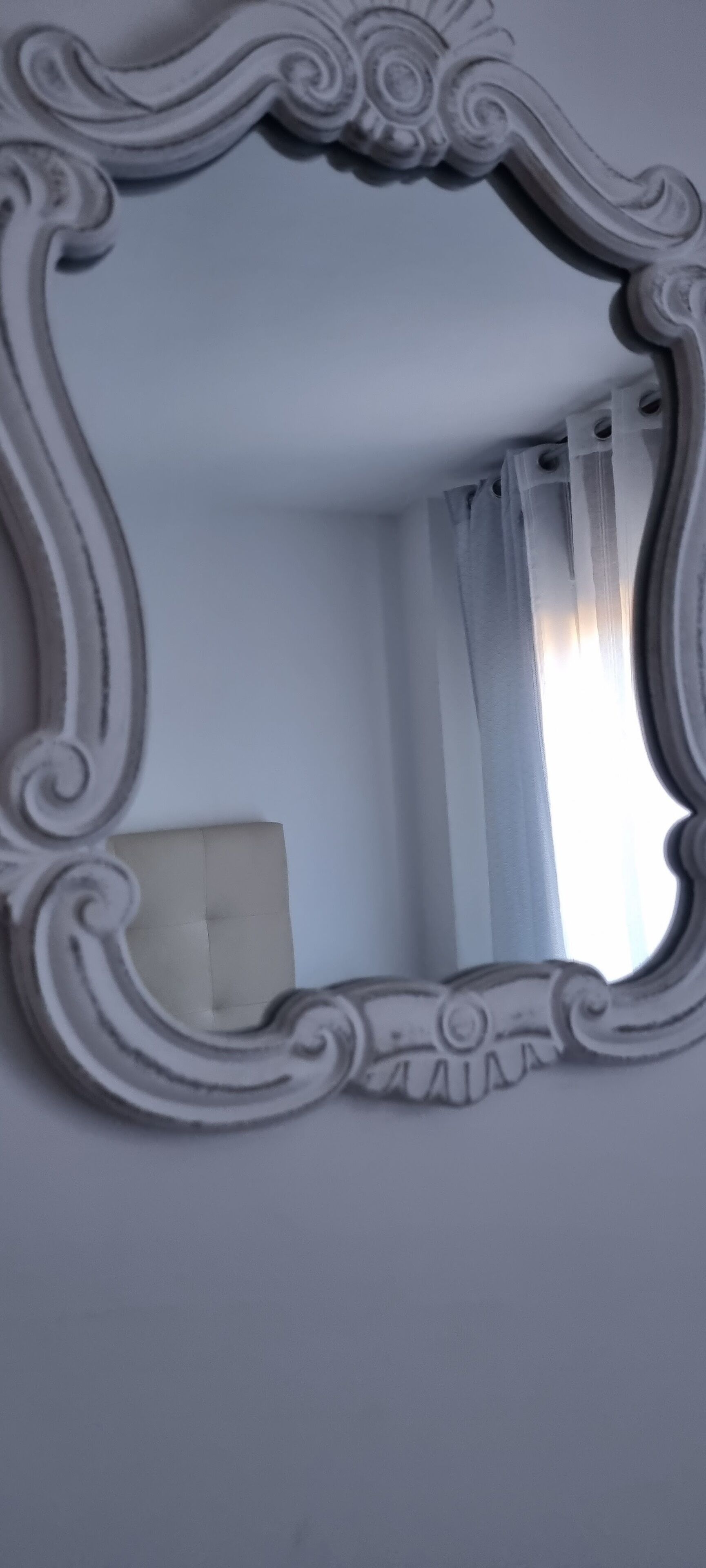 A mirror with ornate, white frame reflecting a room with a white chair and a window with sheer curtains.