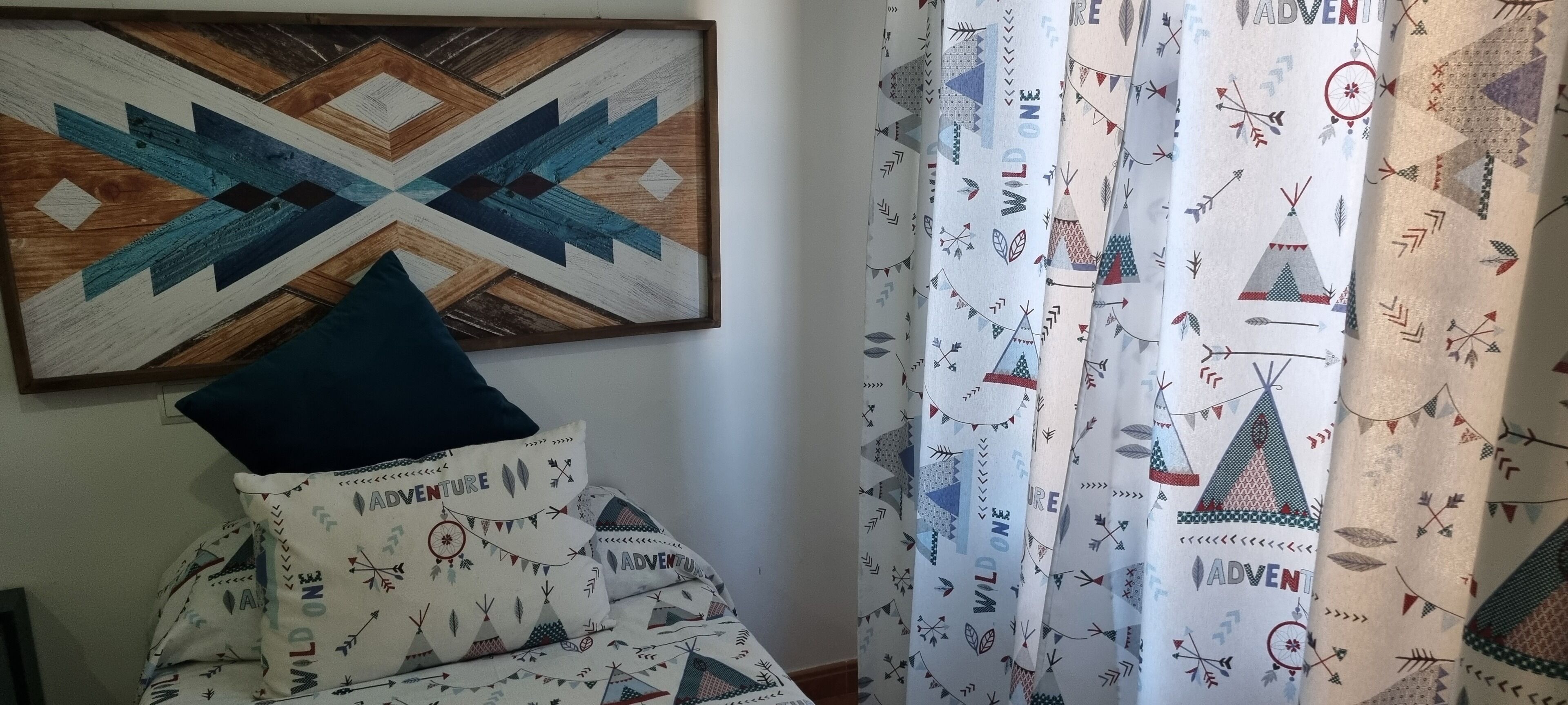 A room with a geometric patterned wall hanging, a bed with a 'WILD ON' themed pillowcase, and patterned curtains with a nature-inspired design.