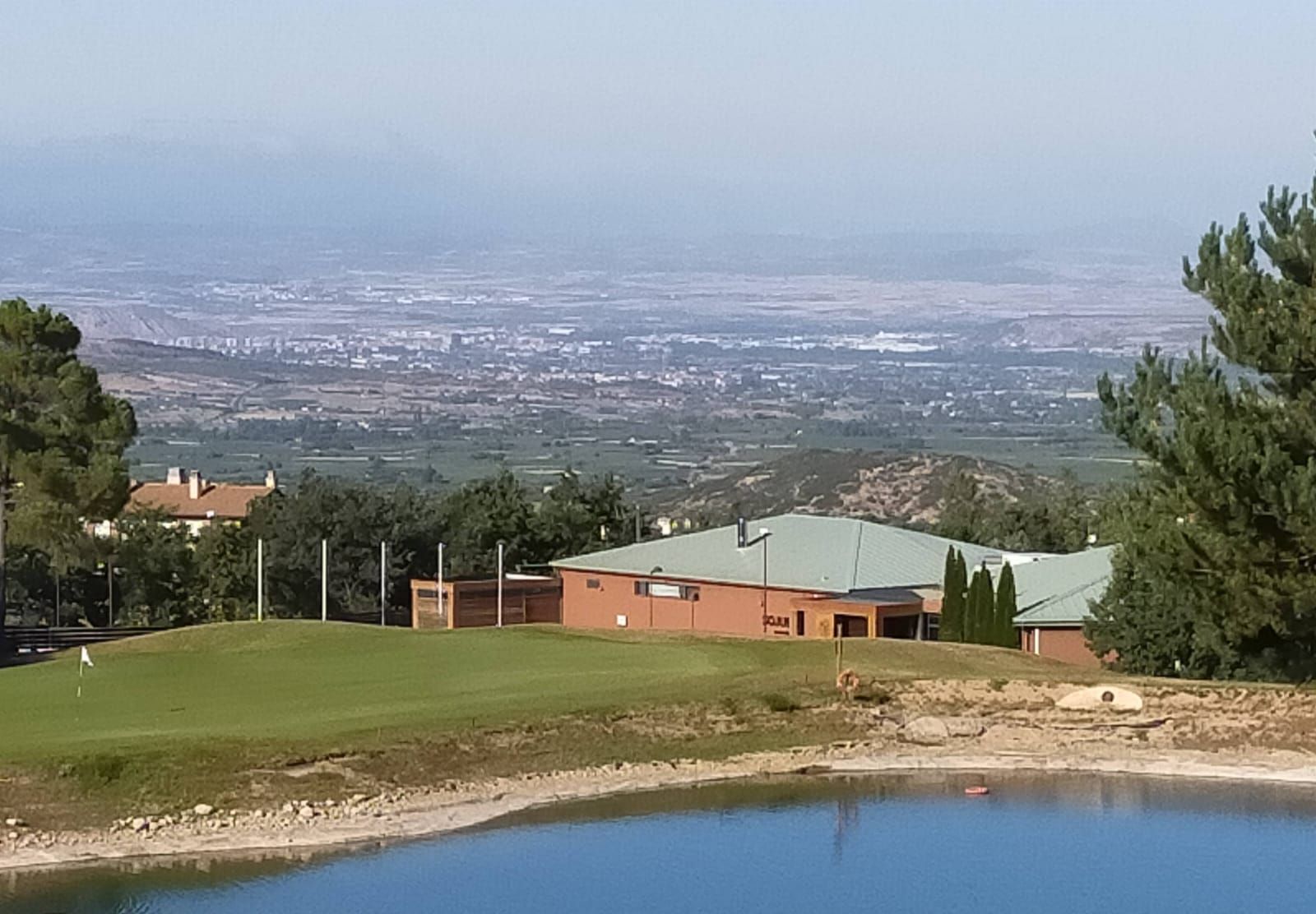Ein Golfplatz mit einem Gebäude, einem Teich und einer entfernten Stadtansicht.