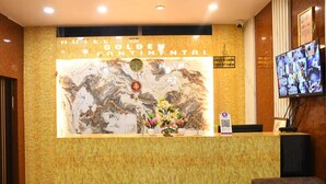 Un mostrador de recepción de hotel con una pared de mármol, un cartel de bienvenida enmarcado, un arreglo floral y un monitor de circuito cerrado.