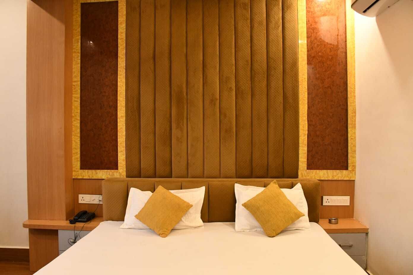 A hotel room with a large bed, two bedside tables, a wooden headboard, and a wall-mounted air conditioning unit.