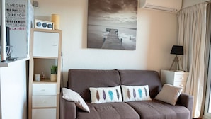 A living room with a brown sofa, a bookshelf, a painting of a pier, a lamp, and a window with curtains. - Alora - Studio Vue Mer Climatisé avec Piscine (Cavalaire-sur-Mer)