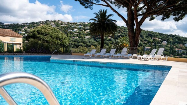 A swimming pool with clear blue water, surrounded by white lounge chairs and a large tree. - Alora - Studio Vue Mer Climatisé avec Piscine (Cavalaire-sur-Mer)
