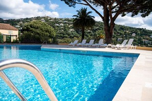 A swimming pool with clear blue water, surrounded by white lounge chairs and a large tree. - Alora - Studio Vue Mer Climatisé avec Piscine (Cavalaire-sur-Mer)