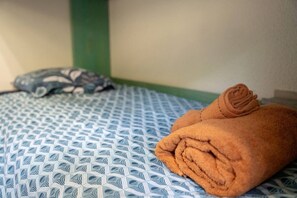 Sängyssä on kuviollinen, sinivalkoinen päiväpeite, taitettu ruskea pyyhe ja harmaa tyyny.