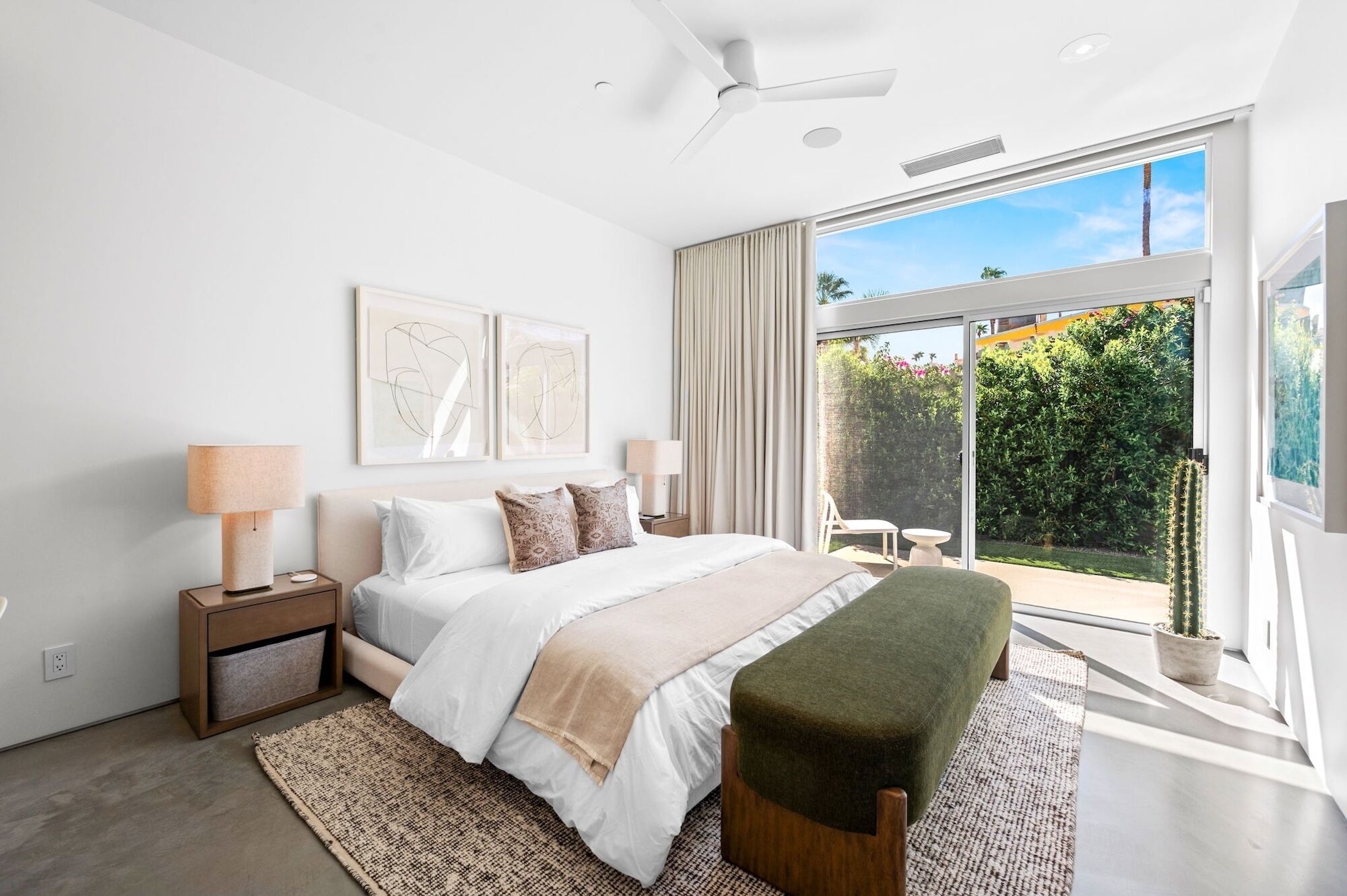 A modern bedroom with a large bed, bedside tables, a bench, and a view of a garden through a sliding glass door.
