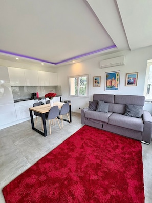 A modern living space with a dining area, a grey sofa, a red rug, and a kitchen in the background.