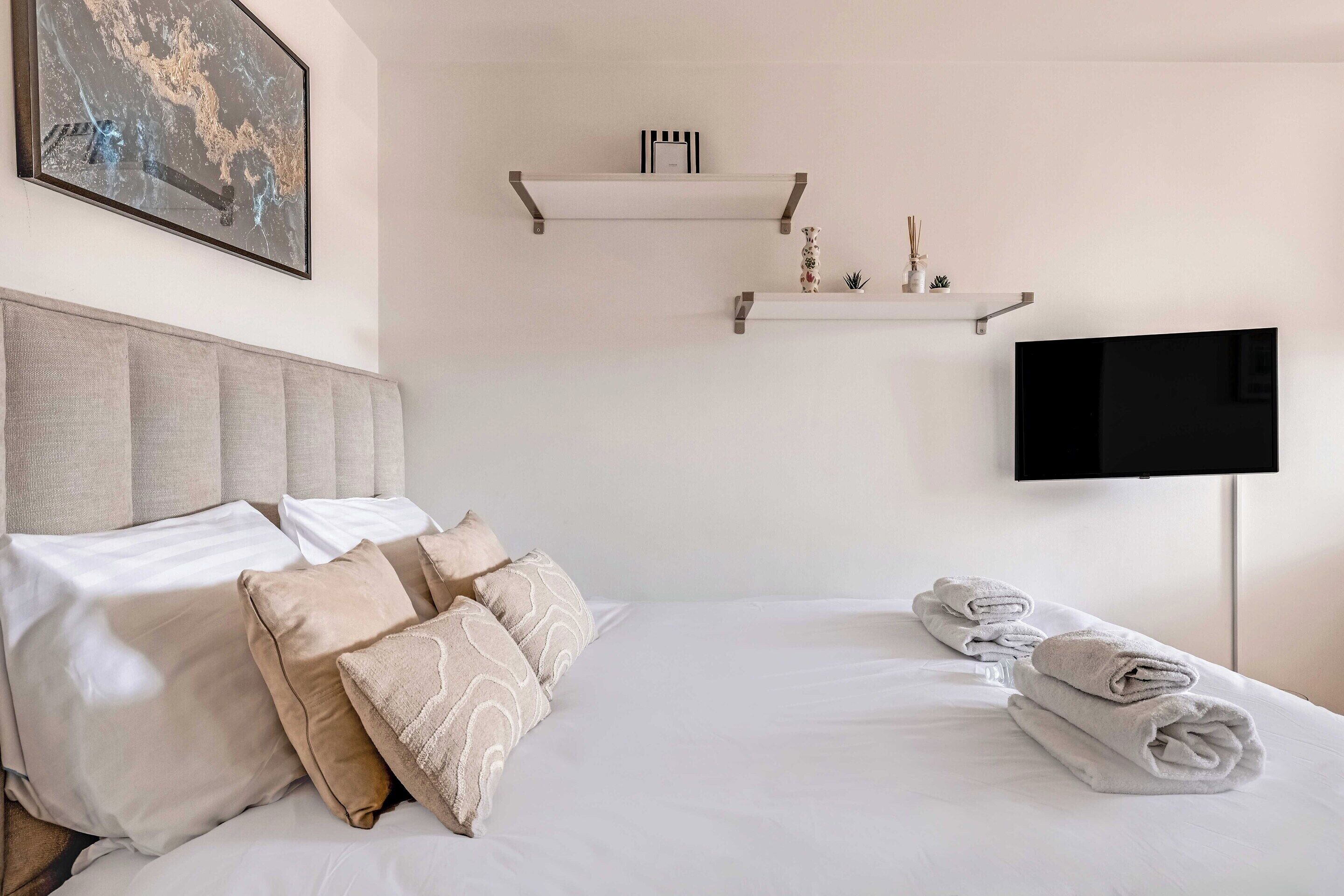 A neatly made bed with pillows, a flat-screen TV mounted on the wall, and two wall-mounted shelves with decorative items.
