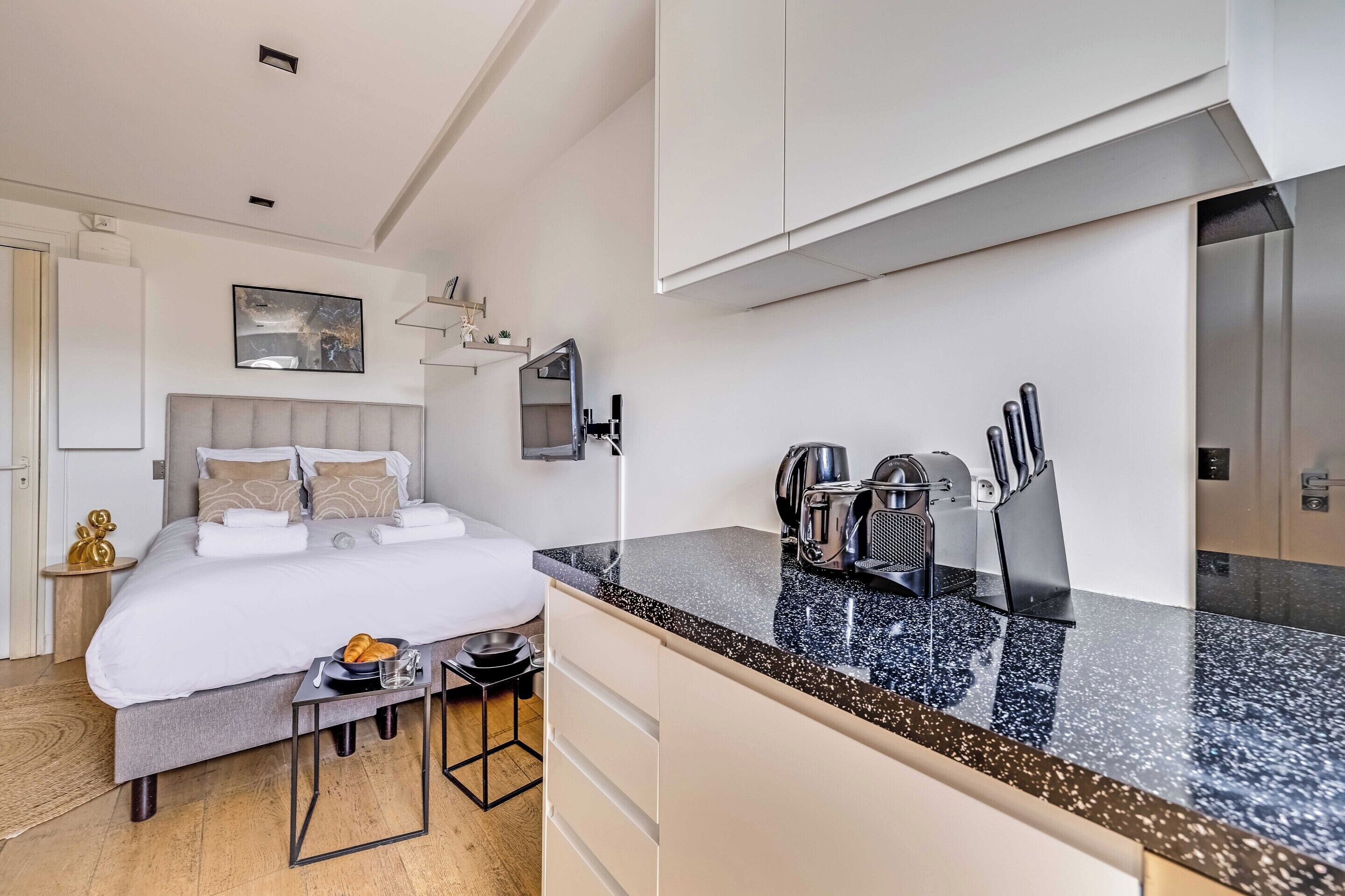 A modern hotel room with a bed, bedside table, and a kitchenette area.