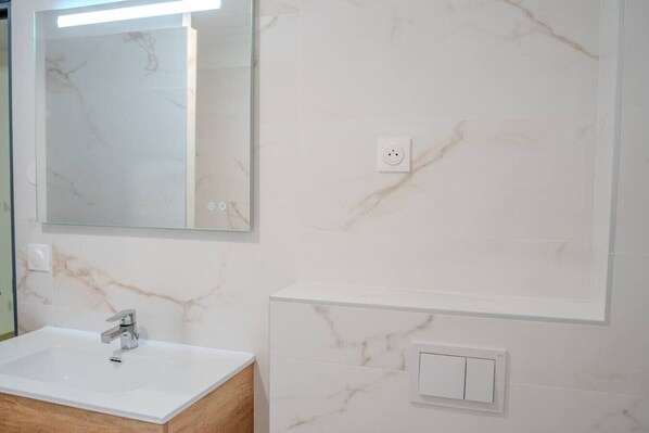 A bathroom with a marble wall, a white sink, a mirror, and a light switch.