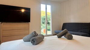 A hotel room with a bed, a flat-screen TV, a dark-colored sofa, and a large window with a view of greenery. - Lou Tavan - Villa 4 Chambres - Piscine & Jacuzzi (Le Lavandou)