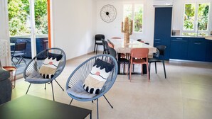 A modern dining area with wicker chairs, a wooden table, and a view of greenery through large windows. - Lou Tavan - Villa 4 Chambres - Piscine & Jacuzzi (Le Lavandou)