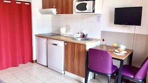 A compact kitchenette with a microwave, sink, and dishwasher, a small dining table with purple chairs, and a TV mounted on the wall.