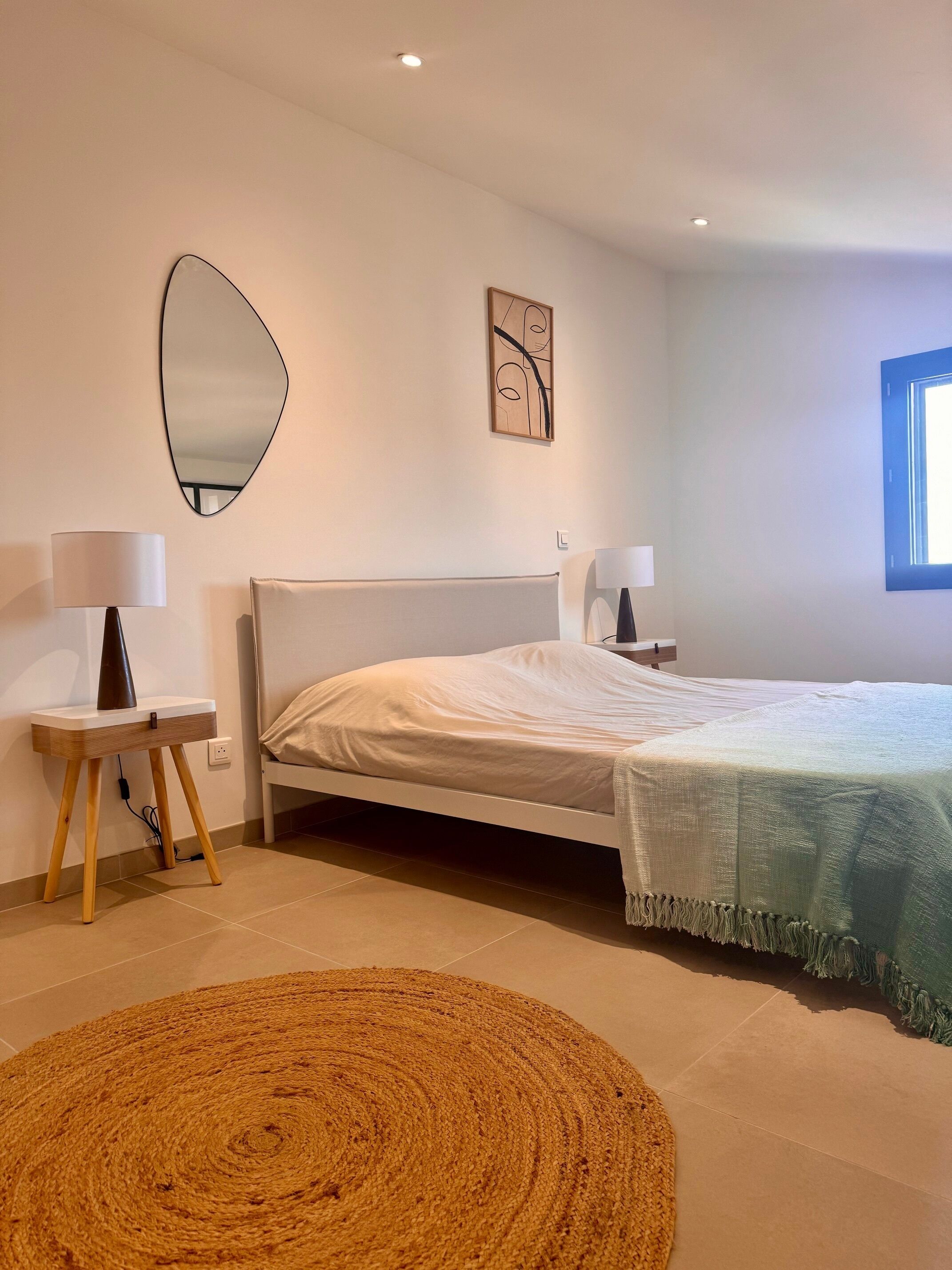 A bedroom with a bed, two bedside tables with lamps, a large mirror, a wall art piece, and a window.