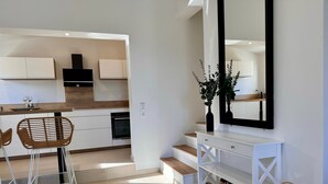 Una cocina moderna con armarios blancos, una zona de comedor con sillas de mimbre y una consola blanca con un espejo y elementos decorativos.