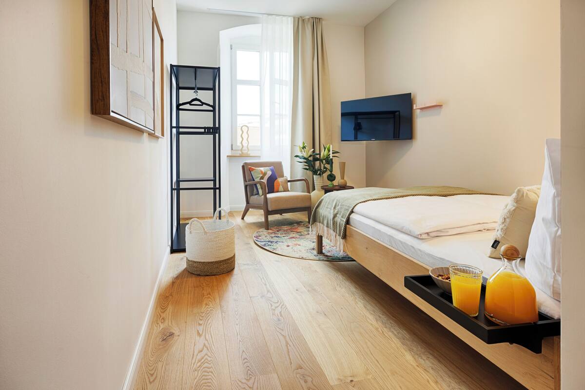 A hotel room with a bed, a bedside table with a tray holding a glass of orange juice and a bottle, a chair, a small table with a vase, and a wall-mounted TV.