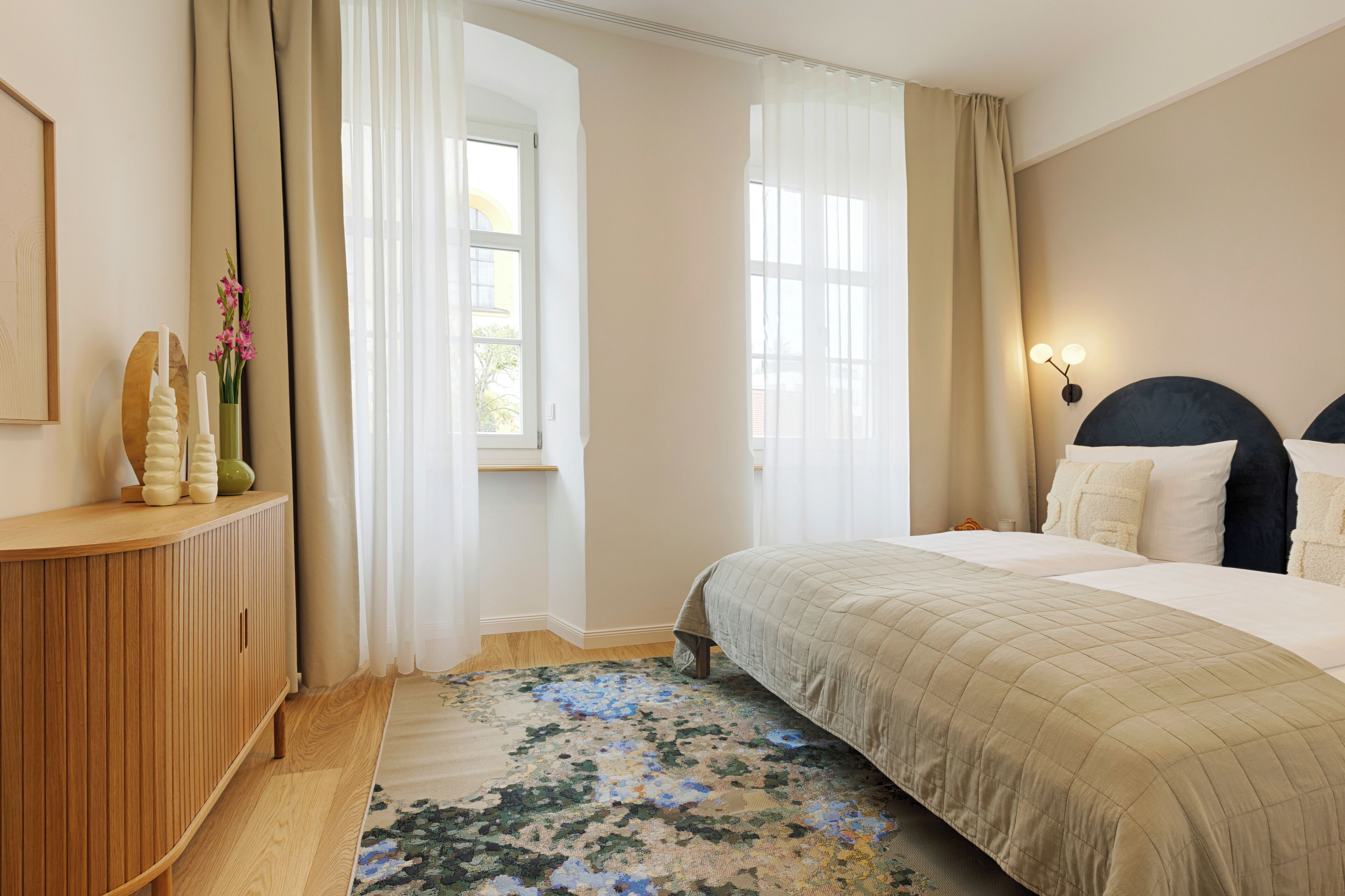 Ein Schlafzimmer mit einem Bett, einer Holzkonsole, einem Fenster mit Vorhängen und einem floralen Teppich.