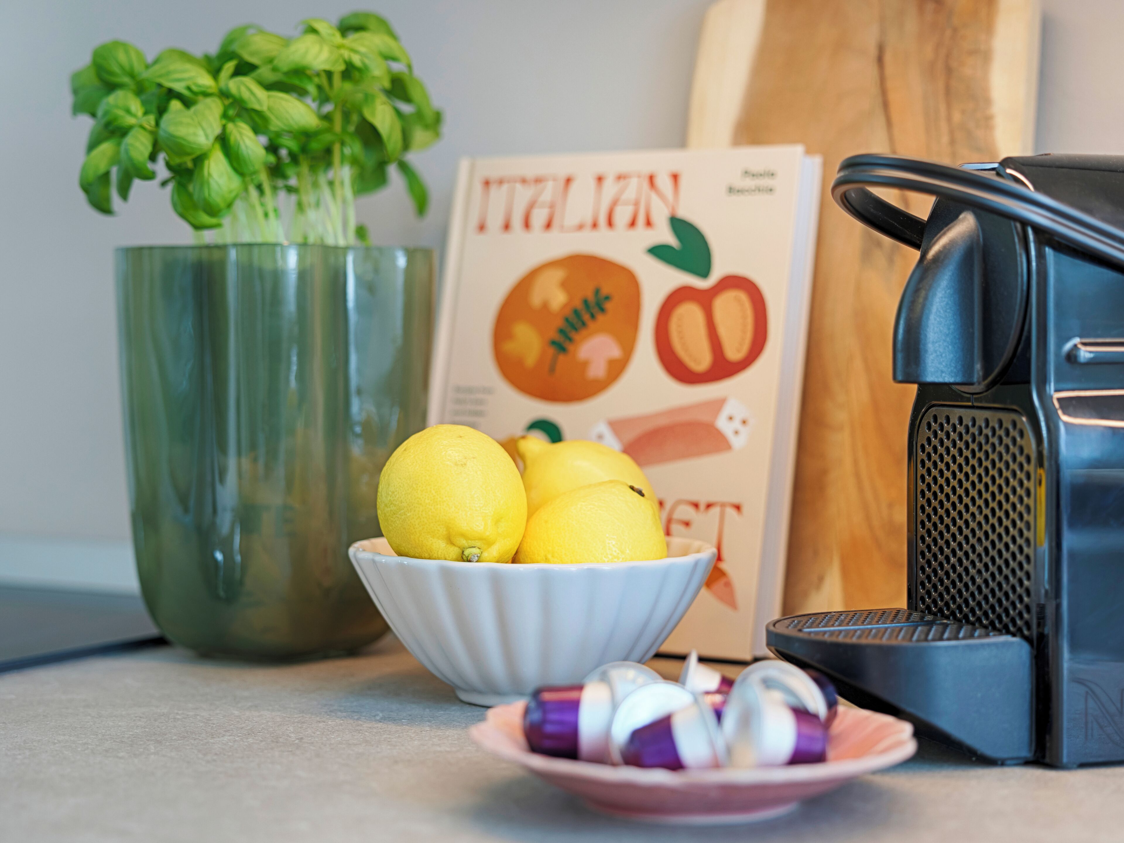 A kitchen counter with a bowl of lemons, a potted basil plant, a cookbook, and a coffee maker.