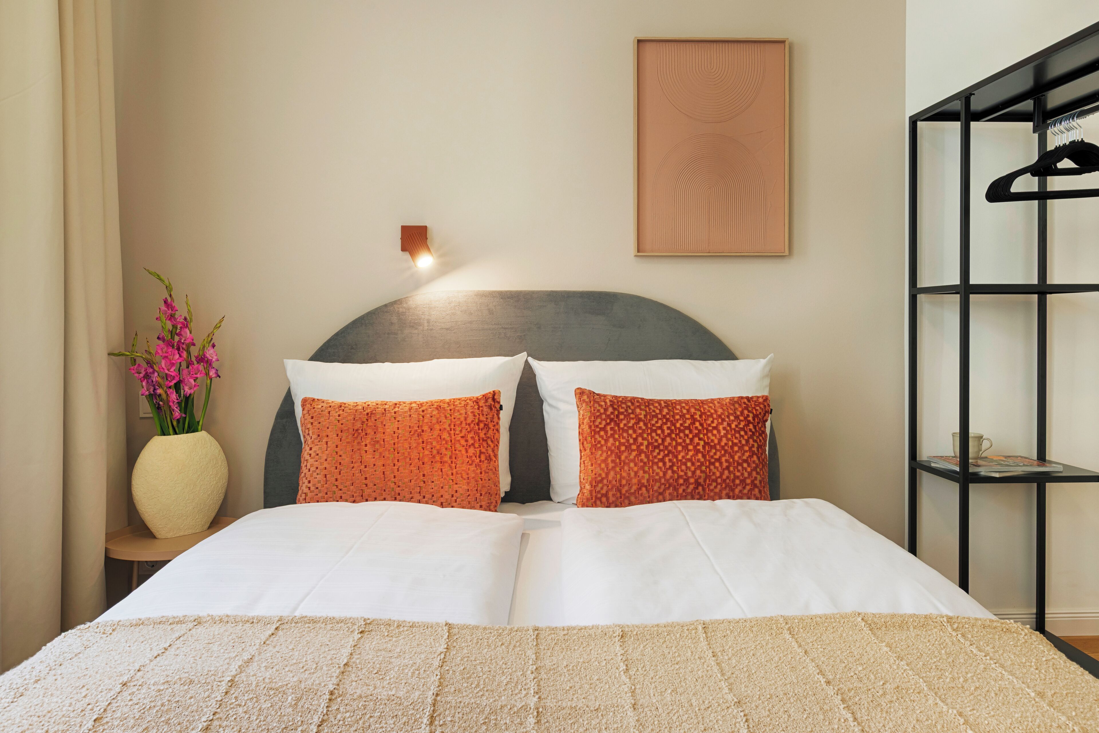 Ein ordentlich bezogenes Bett mit weißen und orangefarbenen Kissen, eine Vase mit rosa Blumen und eine Wandleuchte.