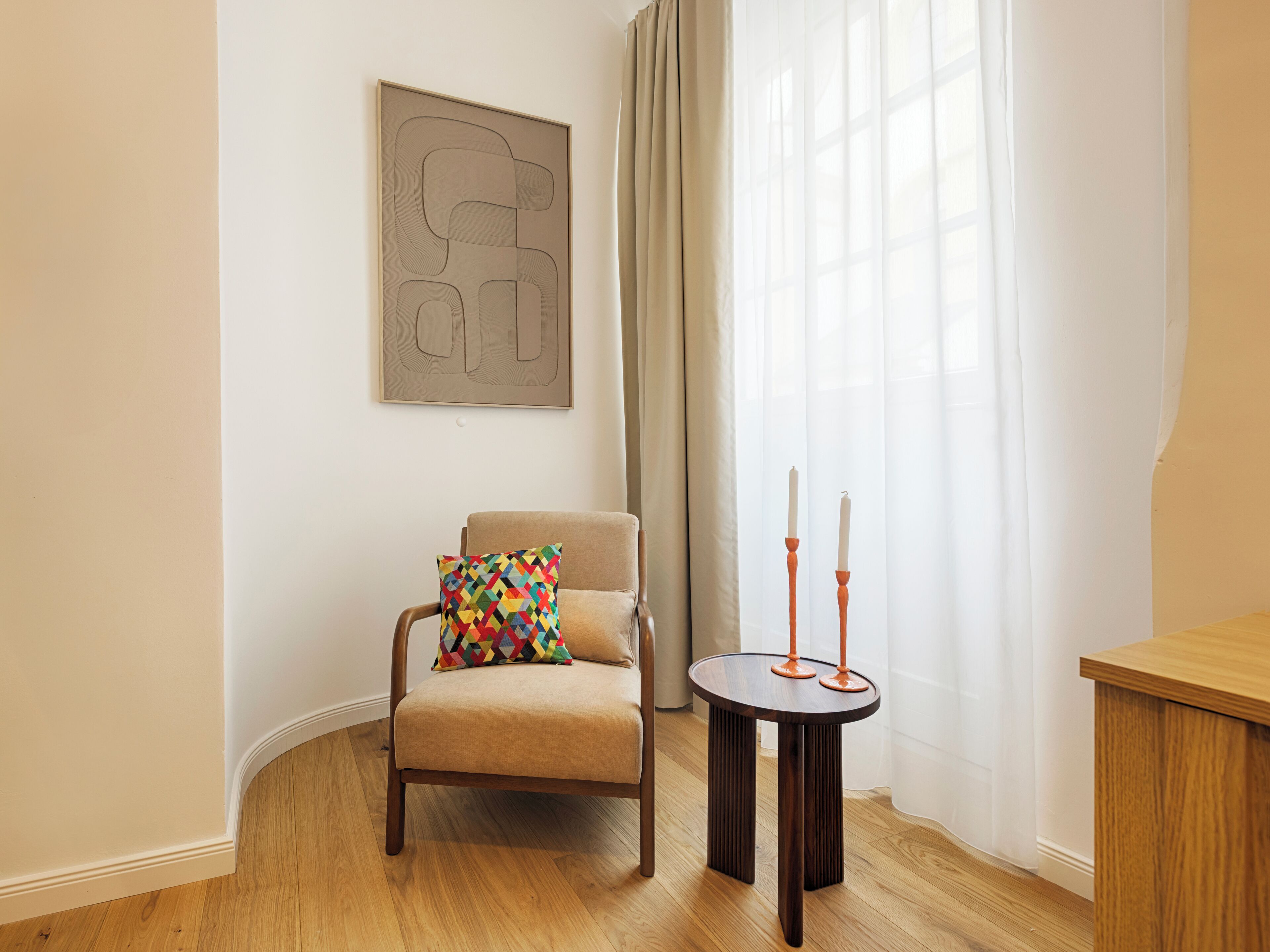A room with a beige armchair, a colorful geometric pillow, a round wooden side table with two candles, and a large abstract painting on the wall.
