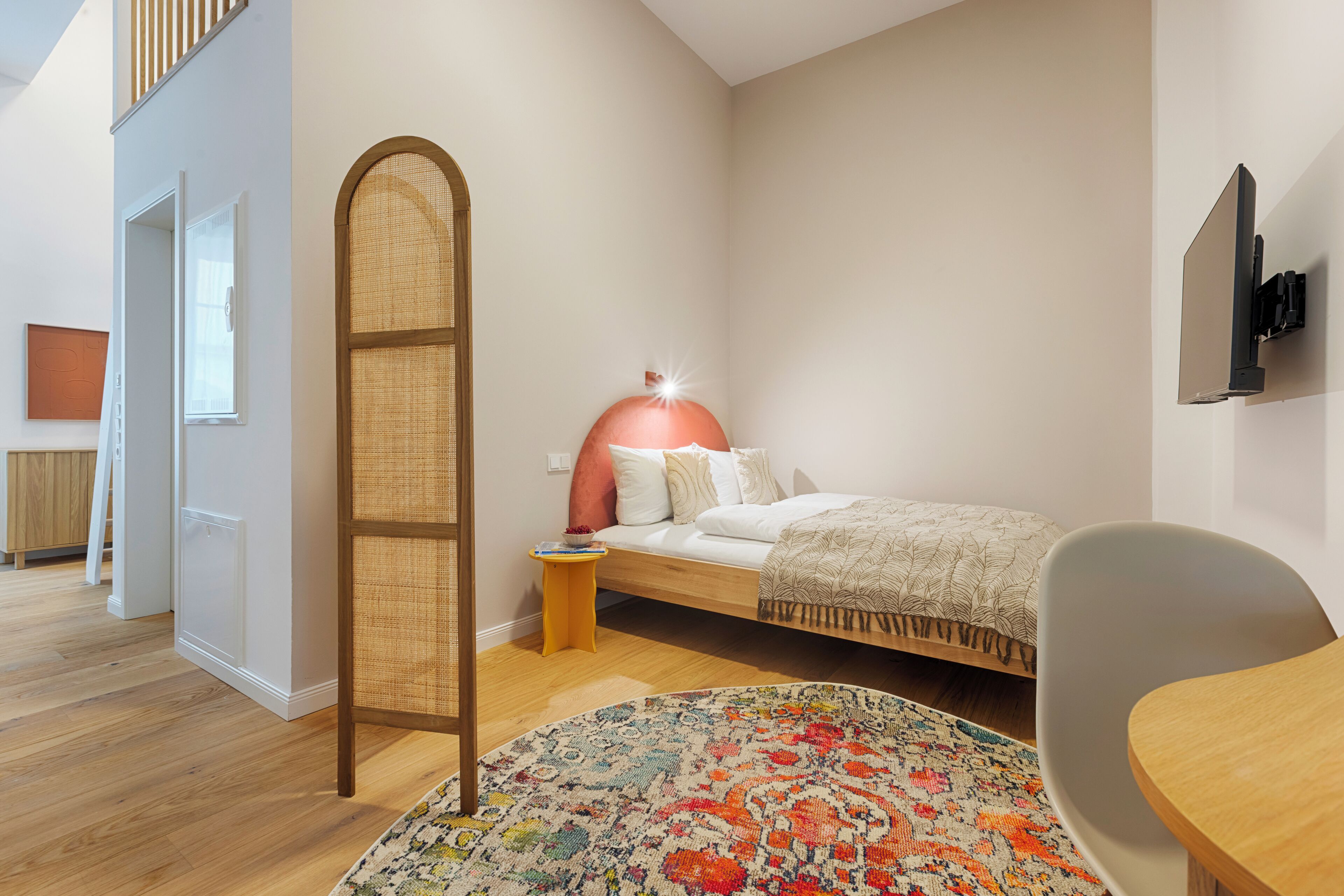 A modern bedroom with a bed, a bedside table, a chair, a colorful rug, and a tall, woven screen.