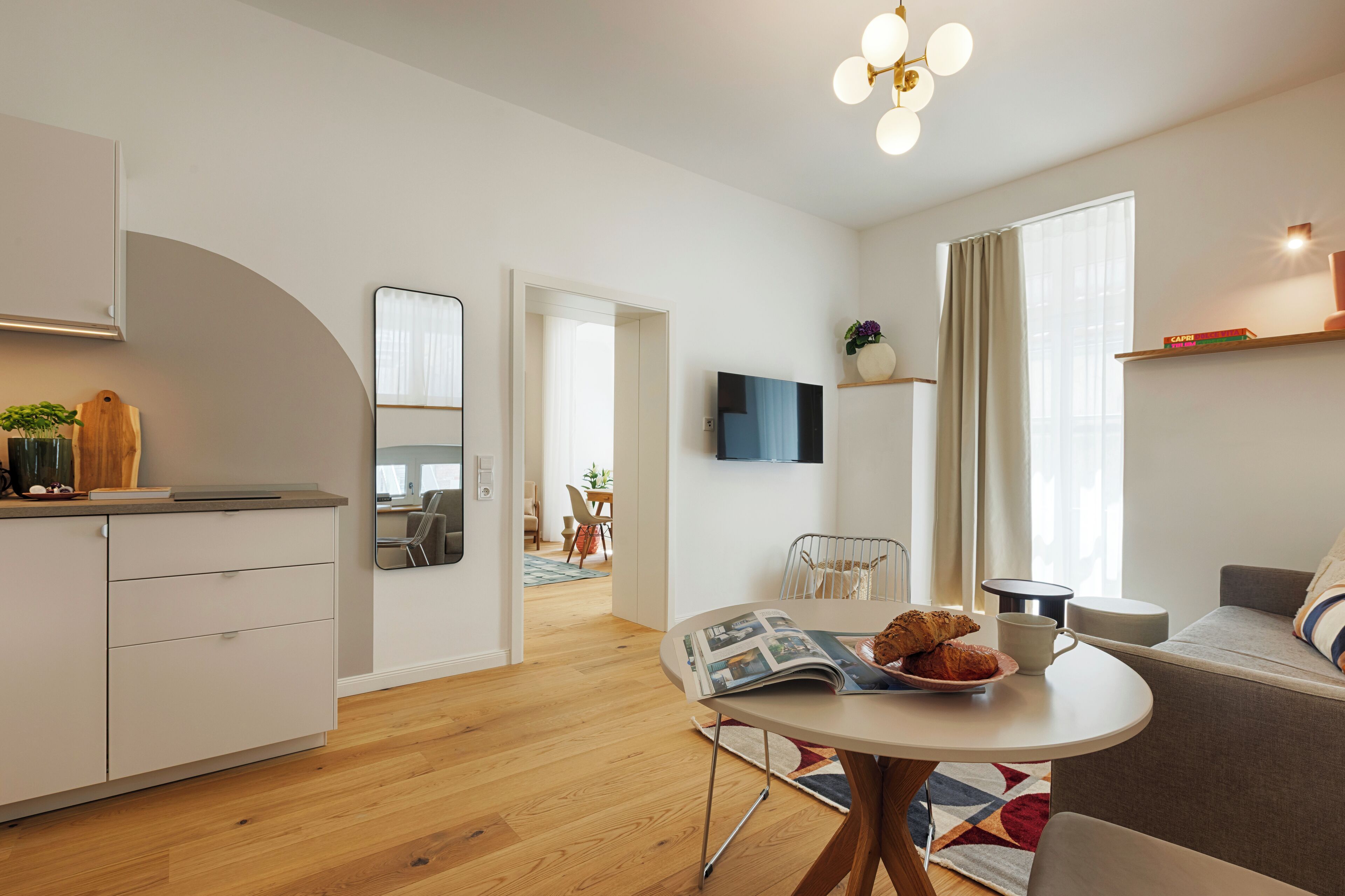 Ein modernes Wohnzimmer mit einer Couch, einem Esstisch mit einer Zeitschrift und Gebäck, einer Küchenzeile mit einer Arbeitsplatte und Schränken sowie einem an der Wand befestigten Fernseher.