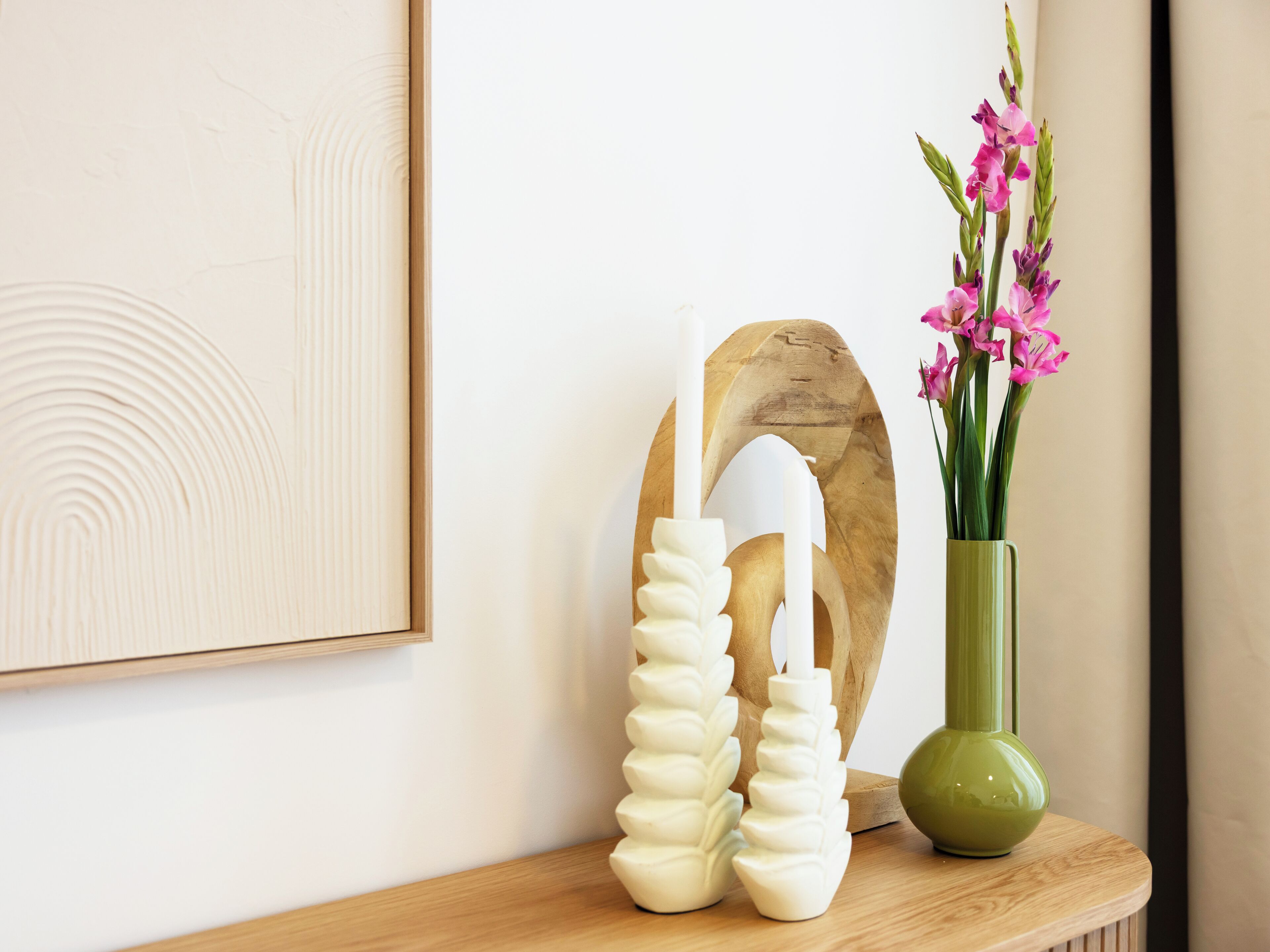 A wooden shelf with a framed artwork, a white abstract sculpture, and a green vase with pink flowers.