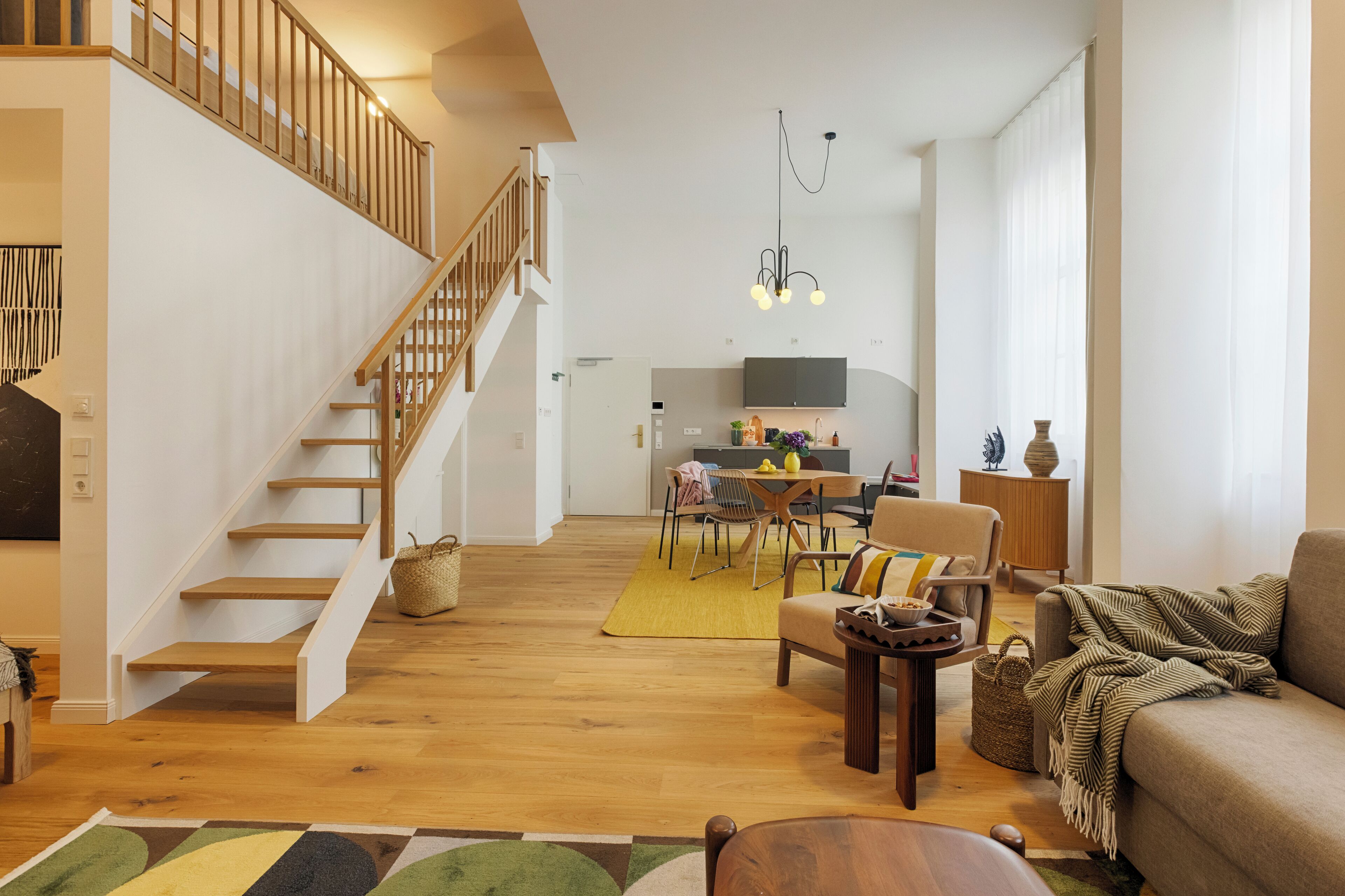 A spacious living area with a wooden staircase, a dining area, and a kitchen in the background.