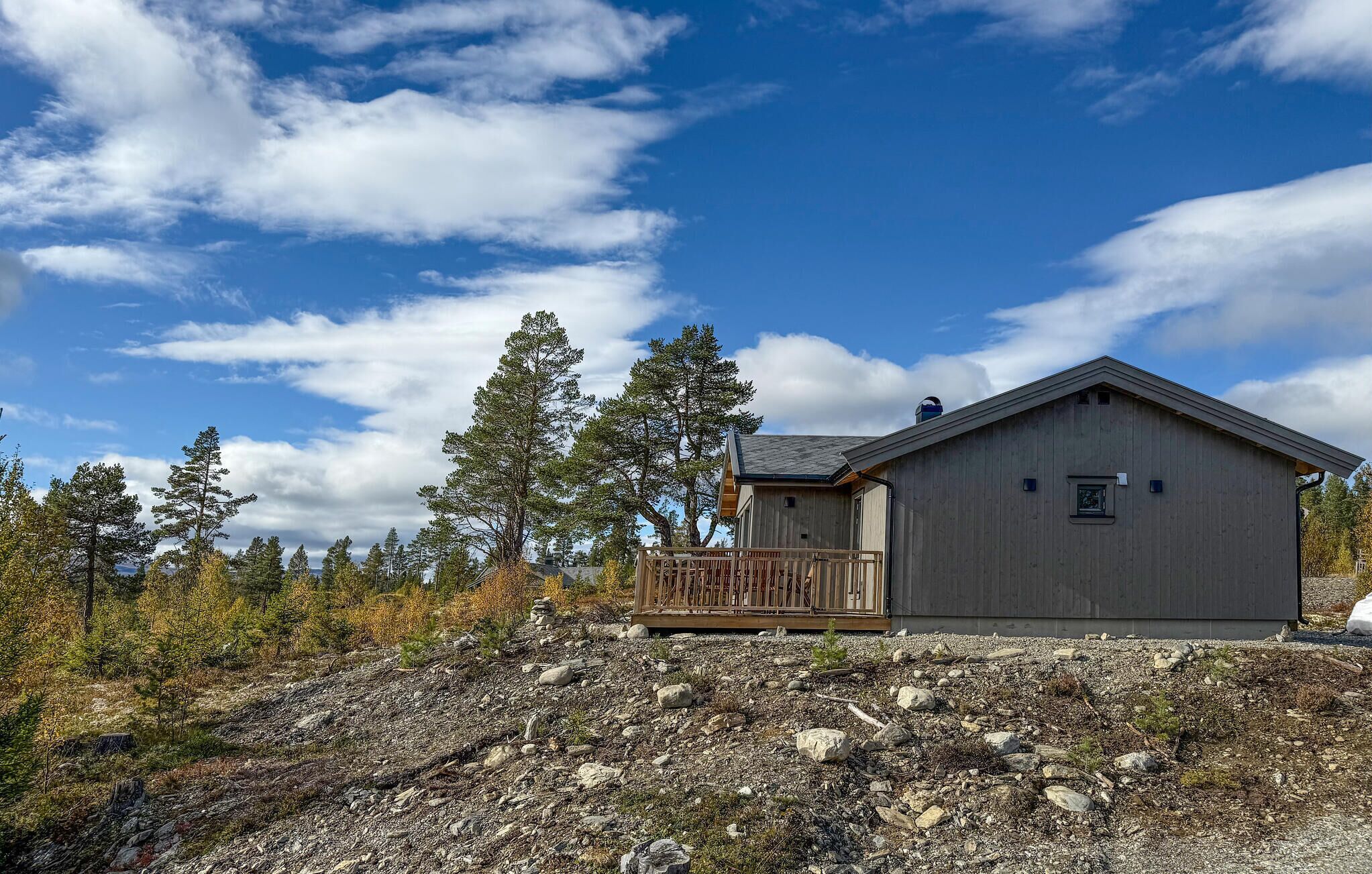 Eine Holzhütte mit Balkon, umgeben von Bäumen und einer felsigen Landschaft unter blauem Himmel.