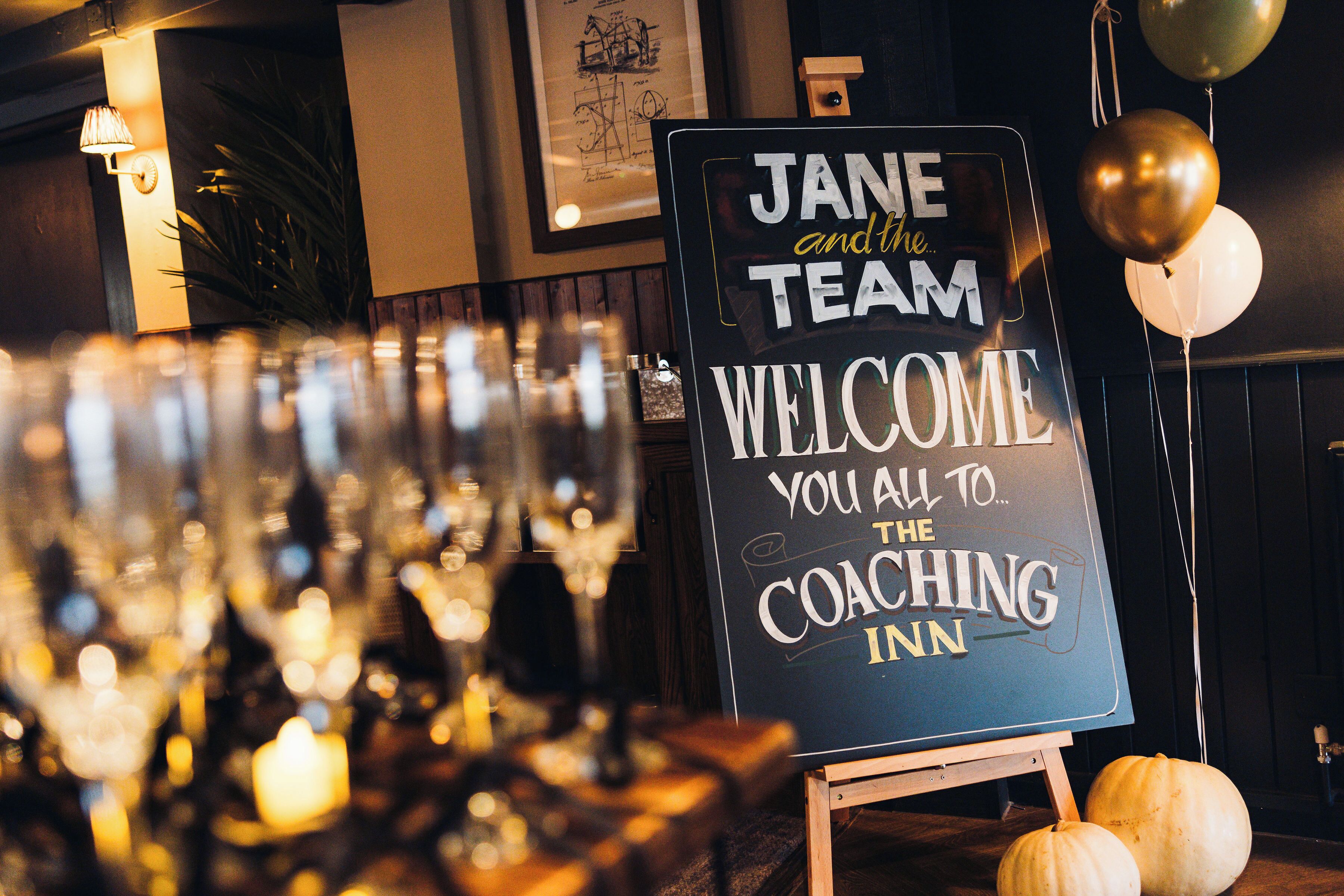 A sign welcoming guests to the Jane and the Team Coaching Inn, with a backdrop of a bar area and decorative pumpkins.