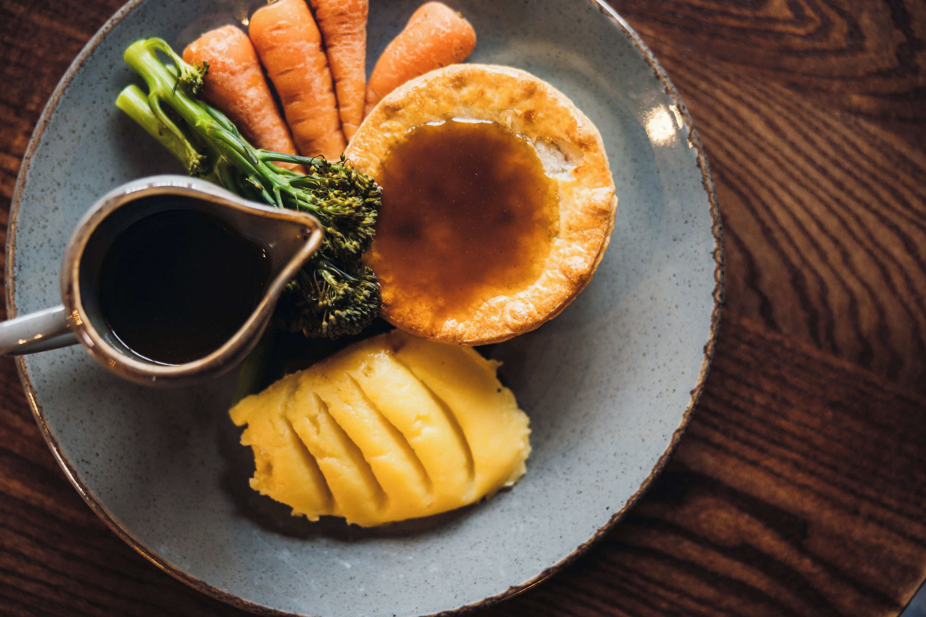Ein Teller mit Yorkshire Pudding, Kartoffelpüree, Karotten und Brokkoli, dazu eine Saucenschale.