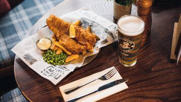 A meal of fish and chips, peas, and sauce on a newspaper, with a beer glass and cutlery on a wooden table.