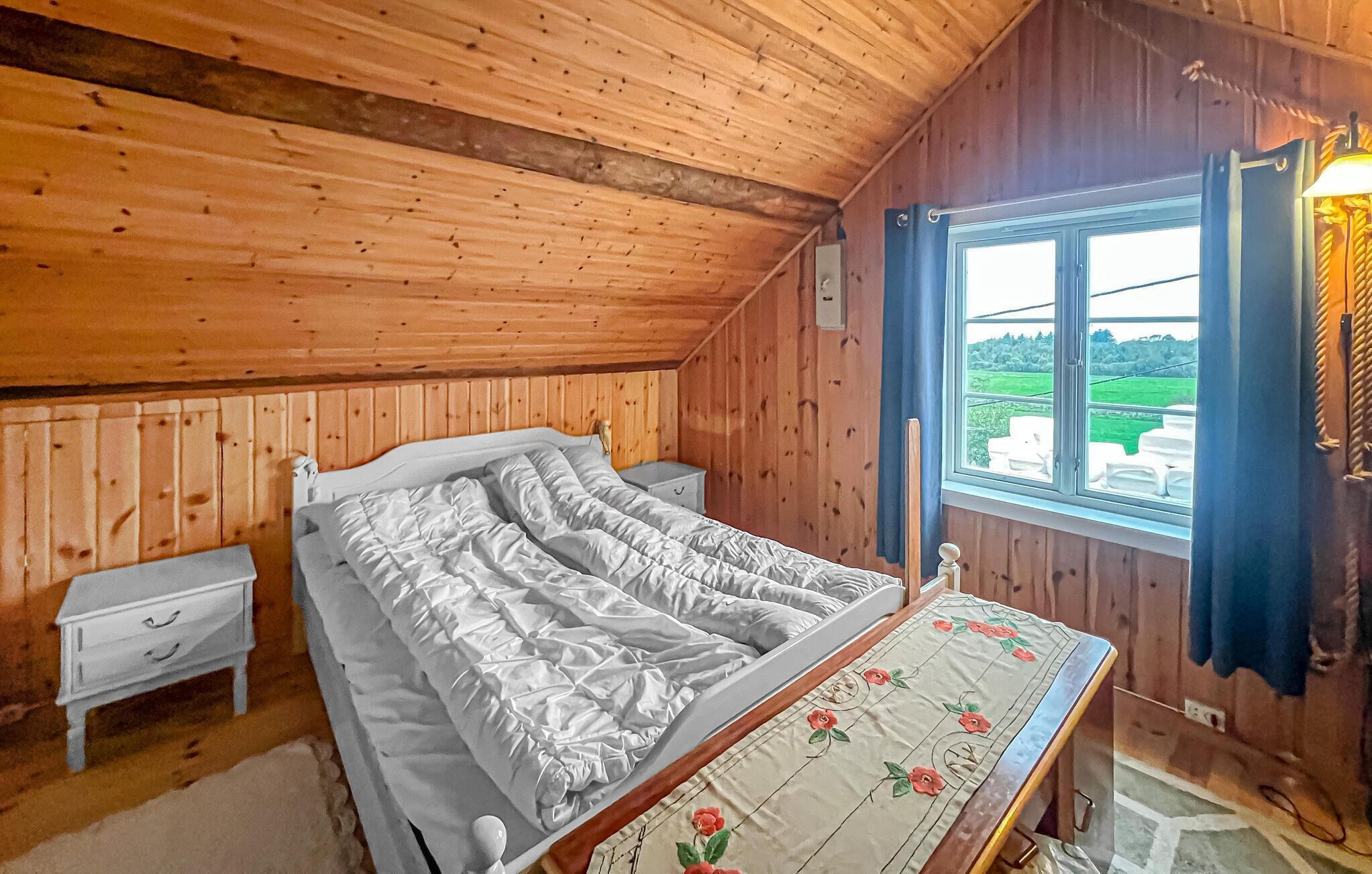 Een slaapkamer met houten lambrisering, een bed, een nachtkastje, een tafel met bloemenonderzetter en een raam met uitzicht op het landschap.