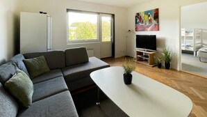 Un salon moderne avec un canapé d’angle gris, une table basse blanche, un téléviseur à écran plat posé sur un meuble en bois et une grande fenêtre donnant sur un paysage arboré.