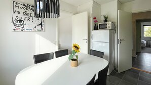 Un coin repas avec une table blanche, des chaises noires, un vase avec des tournesols, et une cuisine équipée d’un four à micro-ondes et d’un réfrigérateur.