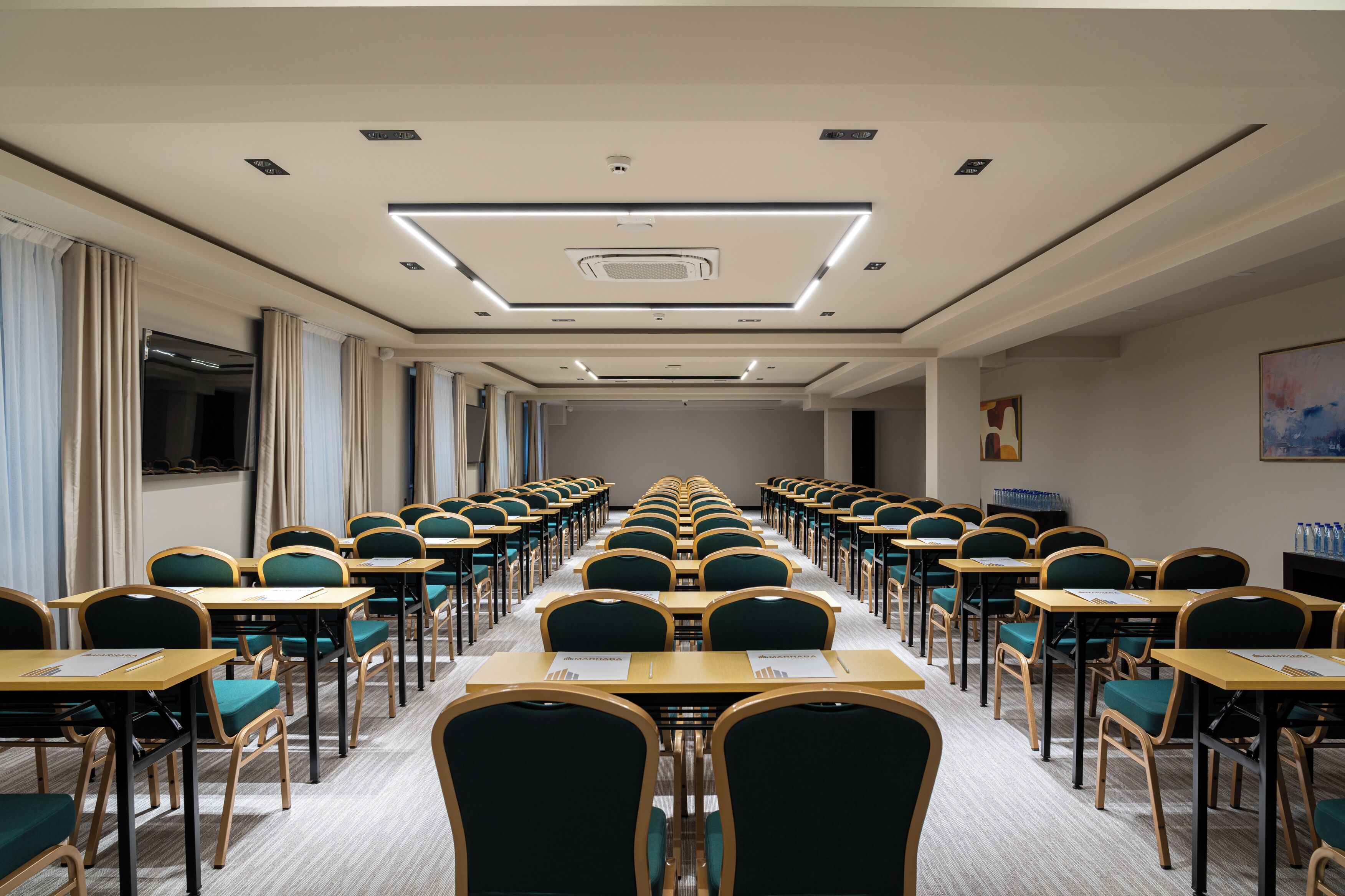 Une salle de conférence avec des rangées de tables et de chaises, un plafond doté d’un éclairage encastré et une grande fenêtre sur le côté gauche.