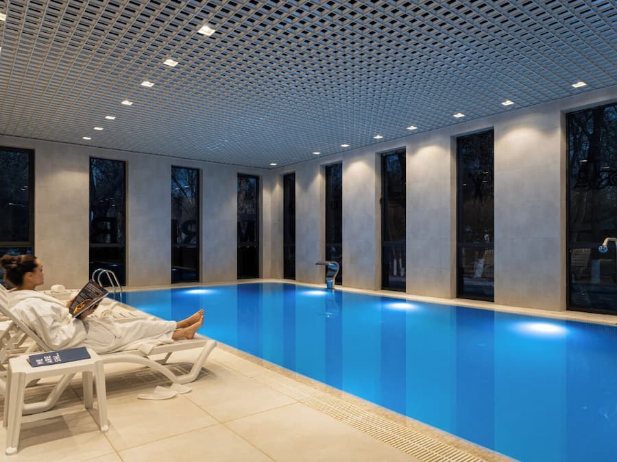 Piscina interior moderna com água azul cristalina, cadeiras de lounge brancas e teto com padrão de grade.